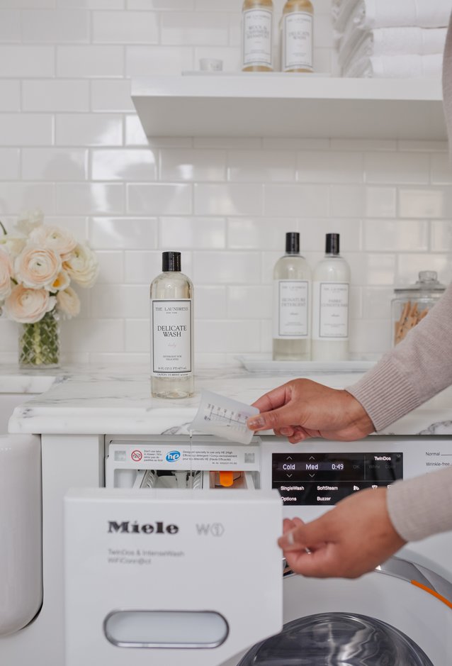 The Laundress Delicate Wash in the scent Lady is poured using The Laundress Measuring Cup into a washing machine.