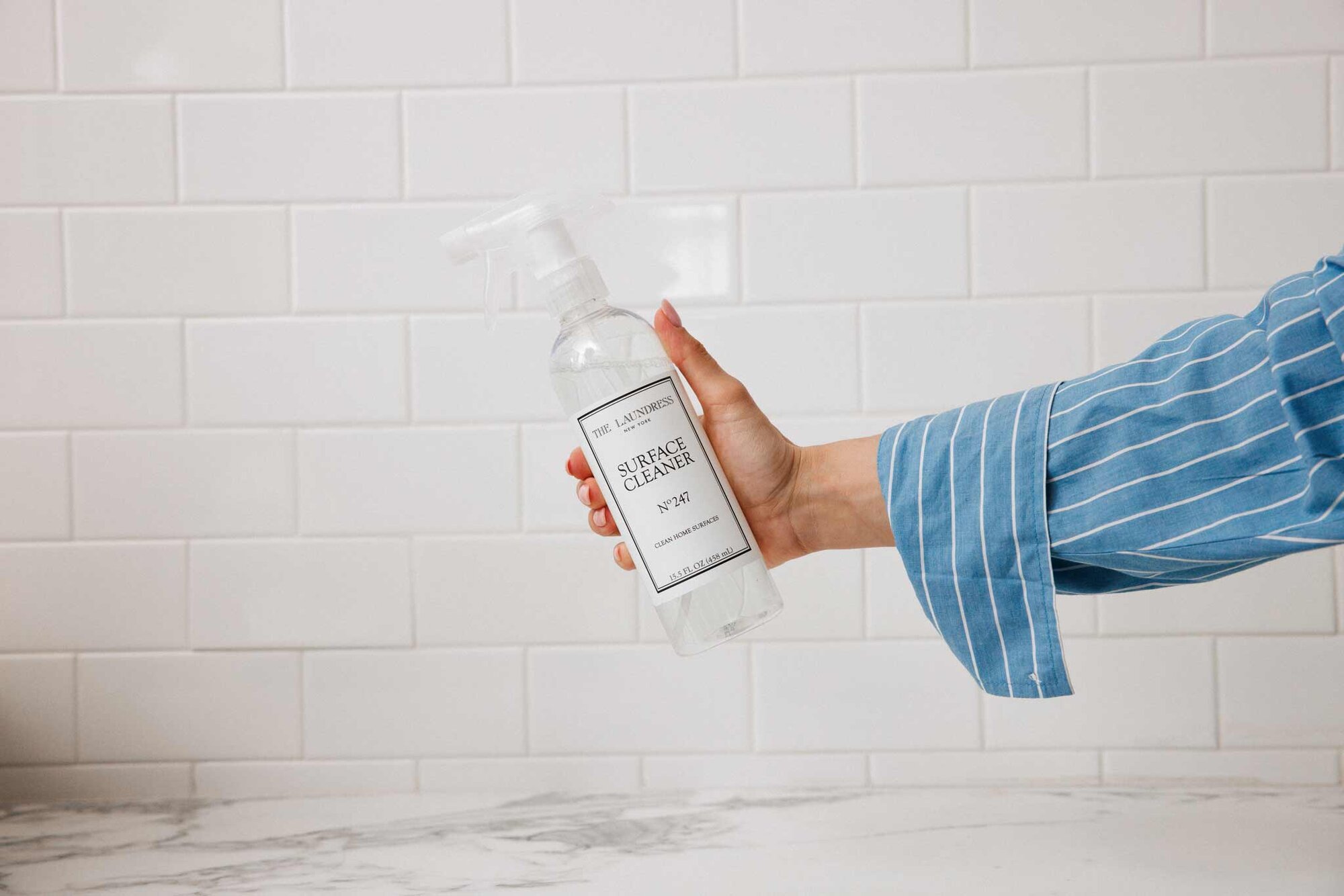 A hand in a striped blue sleeve holds The Laundress Surface Cleaner No. 247 against a white-tiled wall backdrop.