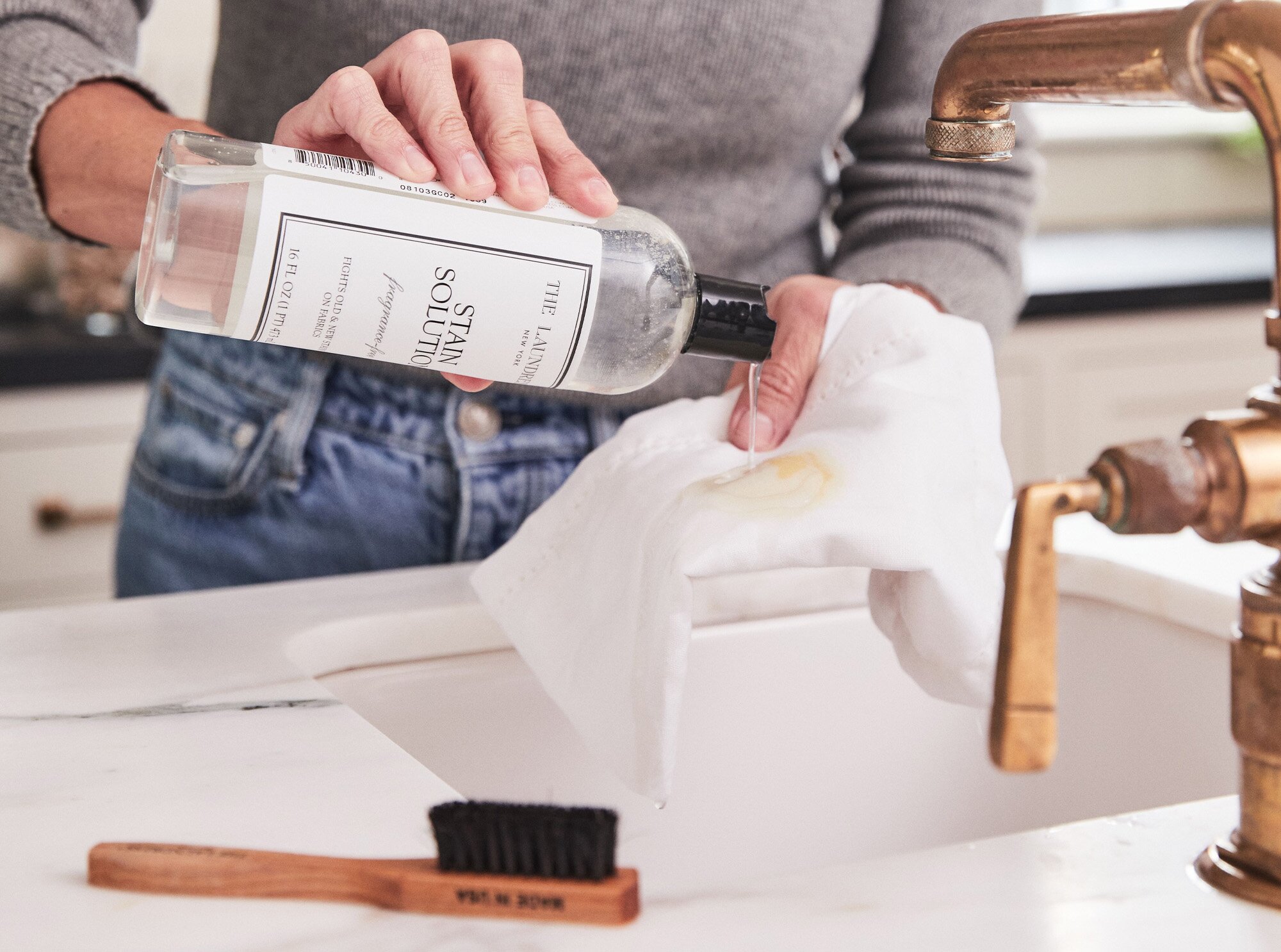 The Laundress Stain Solution being poured onto a stained white cloth over a white sink with a bronze faucet. The Laundress Stain Brush sits next to the sink.