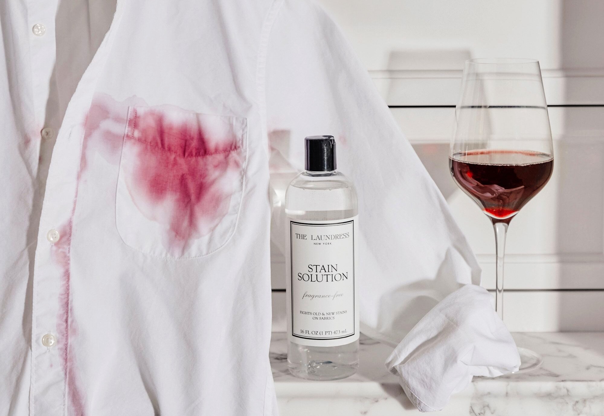 A white shirt stained with red wine next to The Laundress Stain Solution and a half-full wine glass.