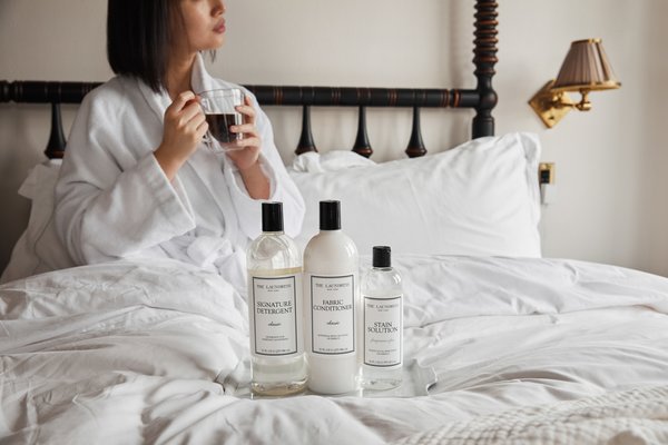 A person in a white bathrobe sits on a bed, holding a mug. The Laundress Signature Detergent, Fabric Conditioner, and Stain Solution are on the bed in front of them.