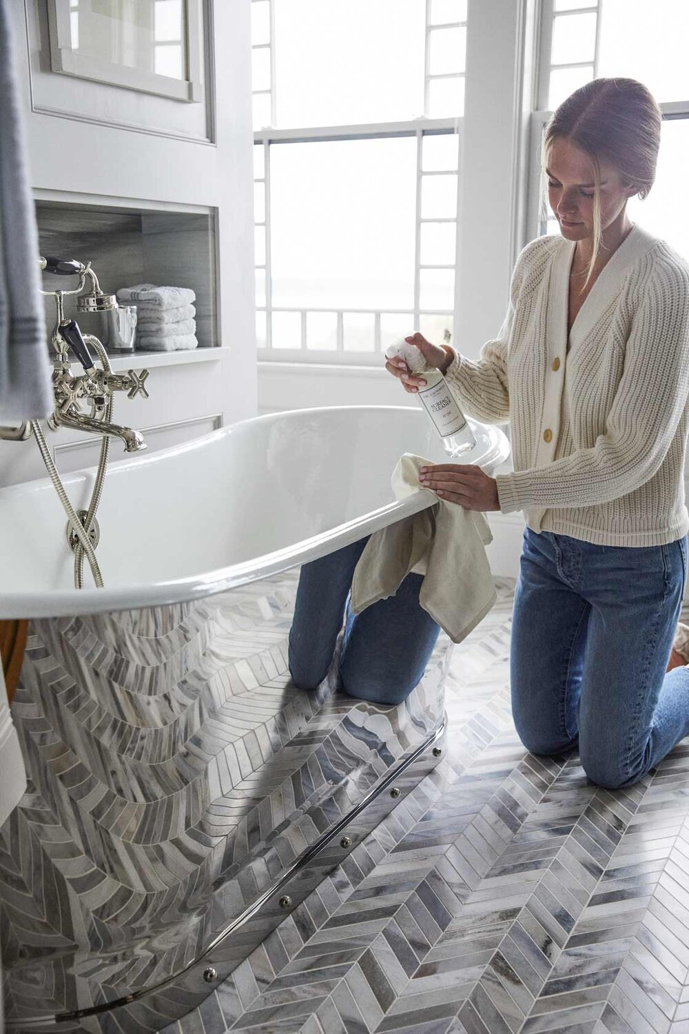 A person kneels beside a white bathtub, spraying The Laundress Surface Cleaner it with a cloth in a bright bathroom featuring patterned marble flooring and large windows.