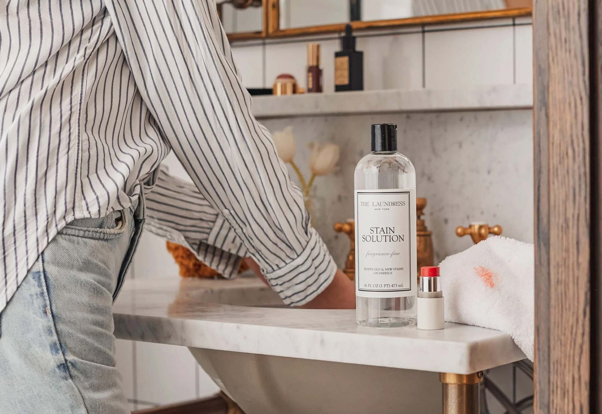 The Laundress Stain Solution sitting on a bathroom countertop beside a lipstick-stained towel and a closed lipstick tube, with a person washing hands under a faucet in a marble sink.