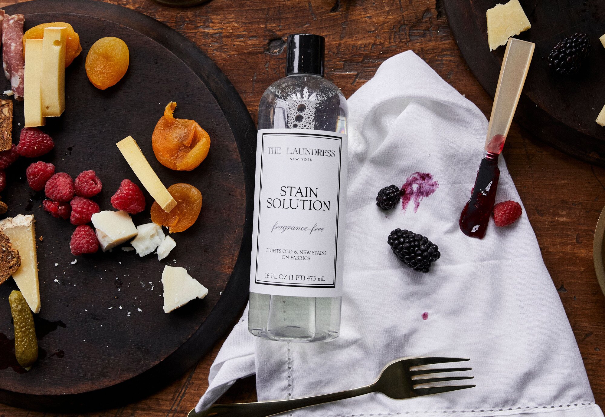 The Laundress Stain Solution on a wooden table next to a food-stained white cloth, surrounded by various fruits, cheese, and a knife.