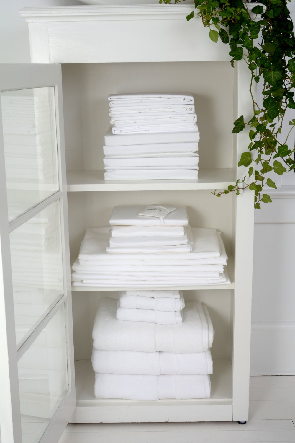 Shelves in a white cabinet holding neatly folded white linens, ready to be stored after being washed with The Laundress's method for cleaning cotton.