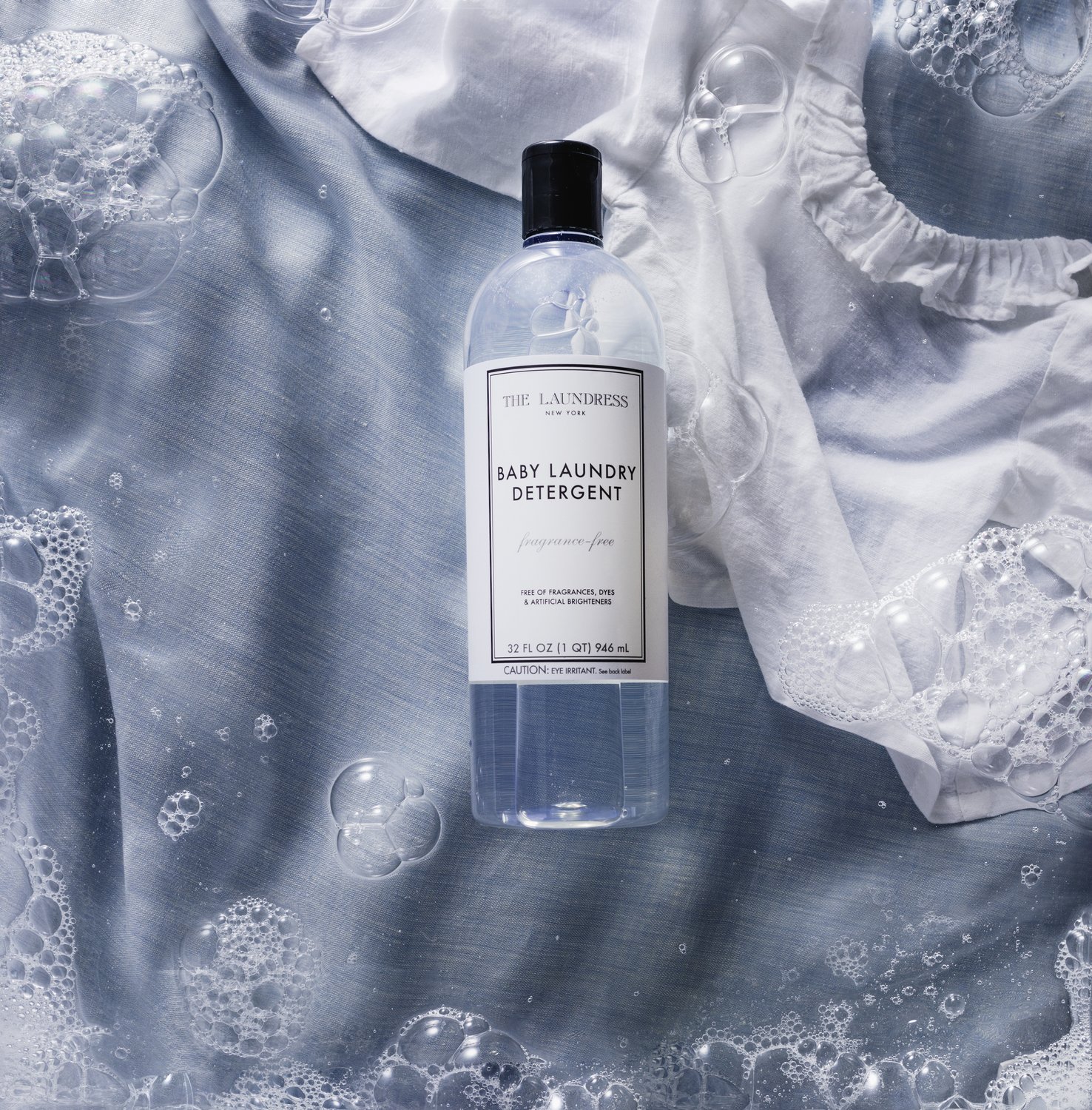 The Laundress Baby Laundry Detergent, free of fragrances, dyes & artificial brighteners, resting on soapy, light blue fabric, alongside white baby clothes.