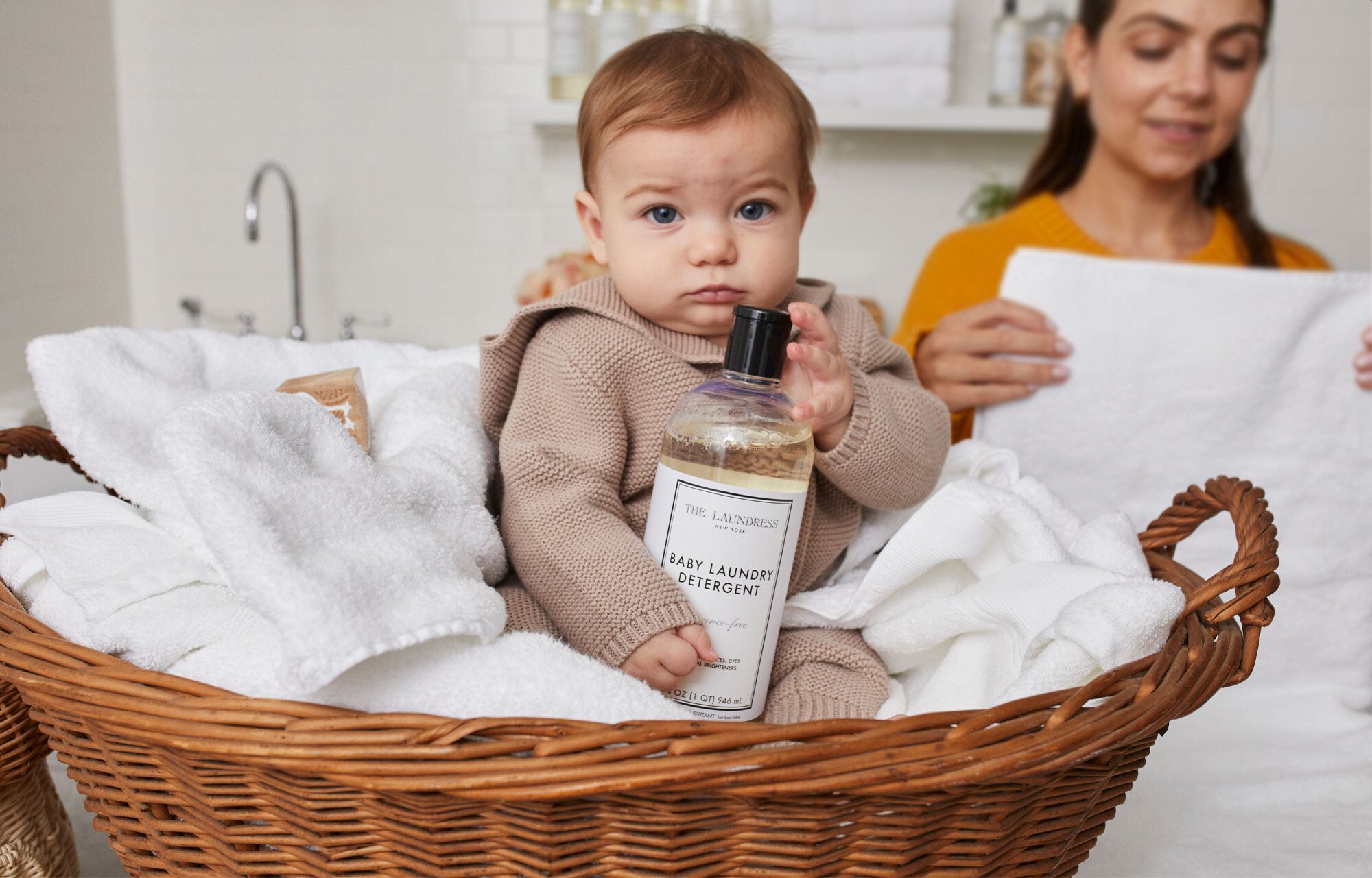 A baby sits in a wicker laundry basket, holding a bottle of The Laundress Baby Laundry Detergent while an adult folds a white towel in the background in a well-lit laundry room.