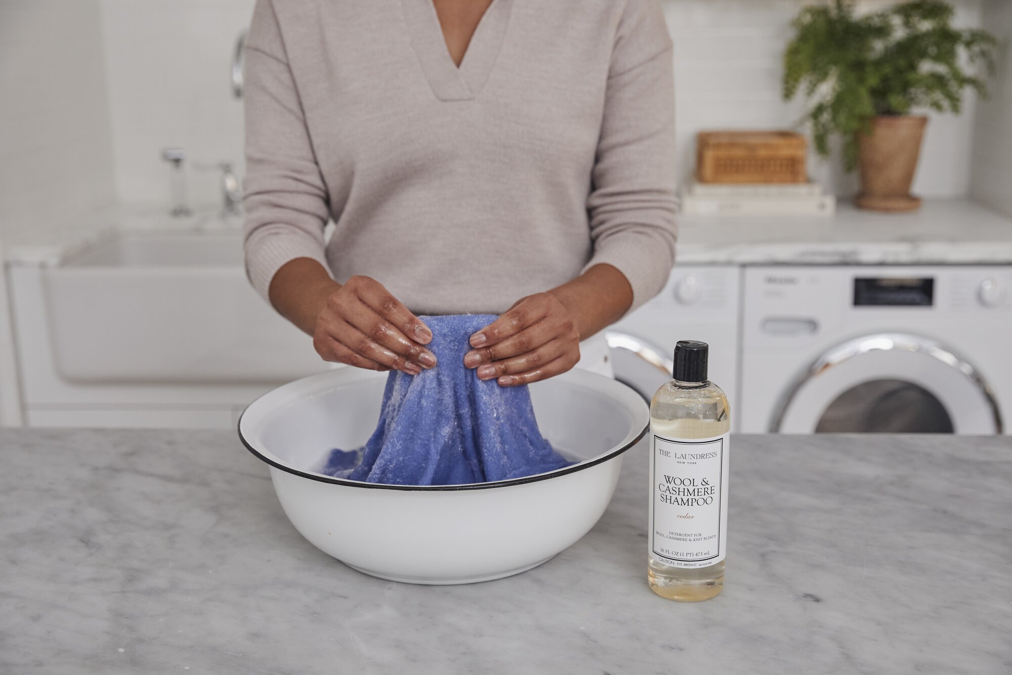 A person washes a blue towel in a white basin using The Laundress Wool & Cashmere Shampoo on a marble countertop, in a laundry room with a washer, dryer, and plants in the background.