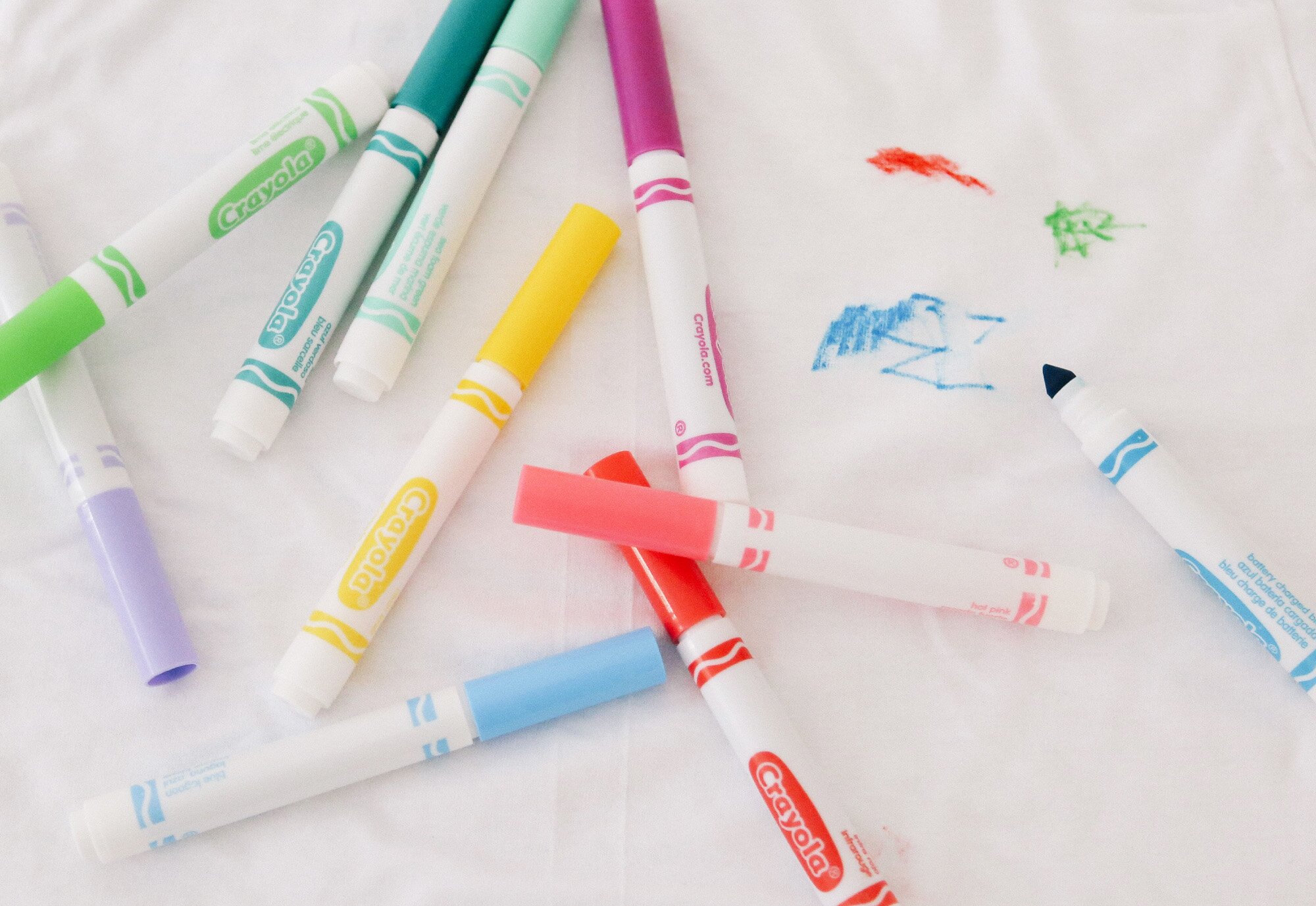 Crayola markers lie scattered on a white fabric, which will be washed using The Laundress' method to removing marker stains from cotton, with a few colorful scribbles (red, green, blue) visible in one corner.