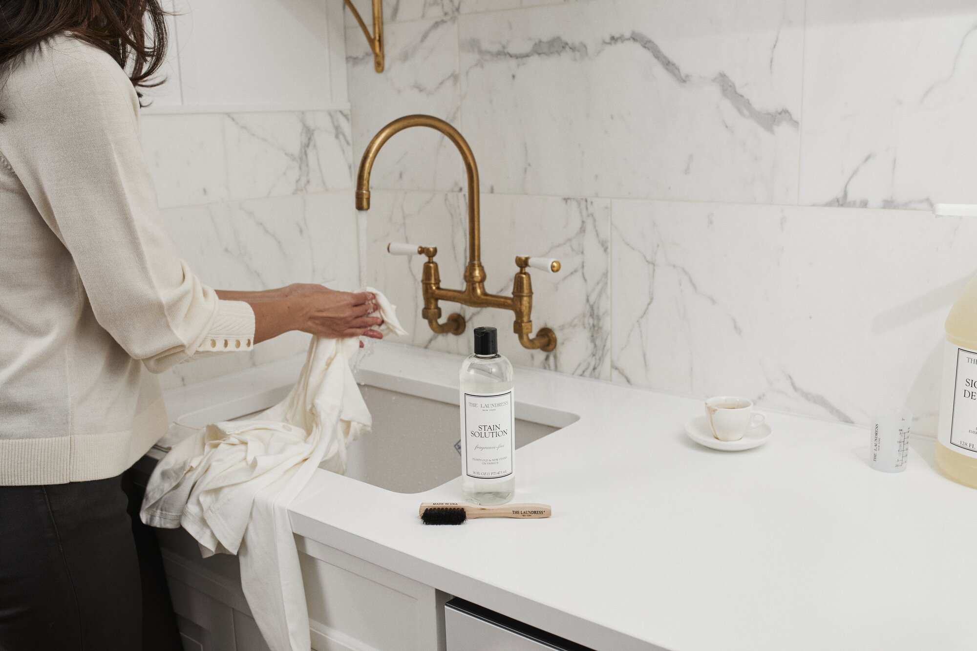 A person washes a white cotton garment under a brass faucet in a white marble-countered kitchen using The Laundress Stain Solution and Stain Brush.