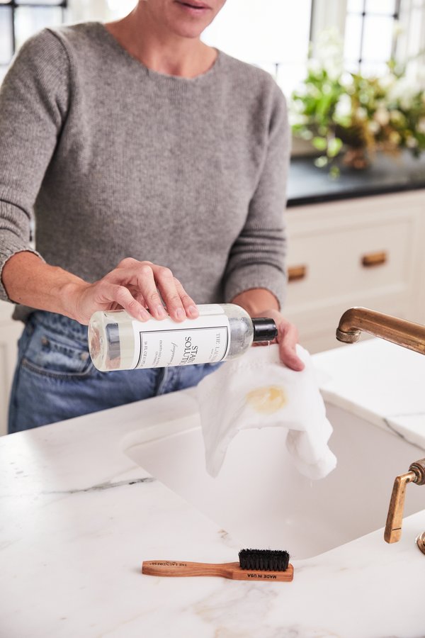 A person wearing a gray sweater pours The Laundress Stain Solution onto a white cloth over a marble sink in a bright kitchen with plants in the background. 