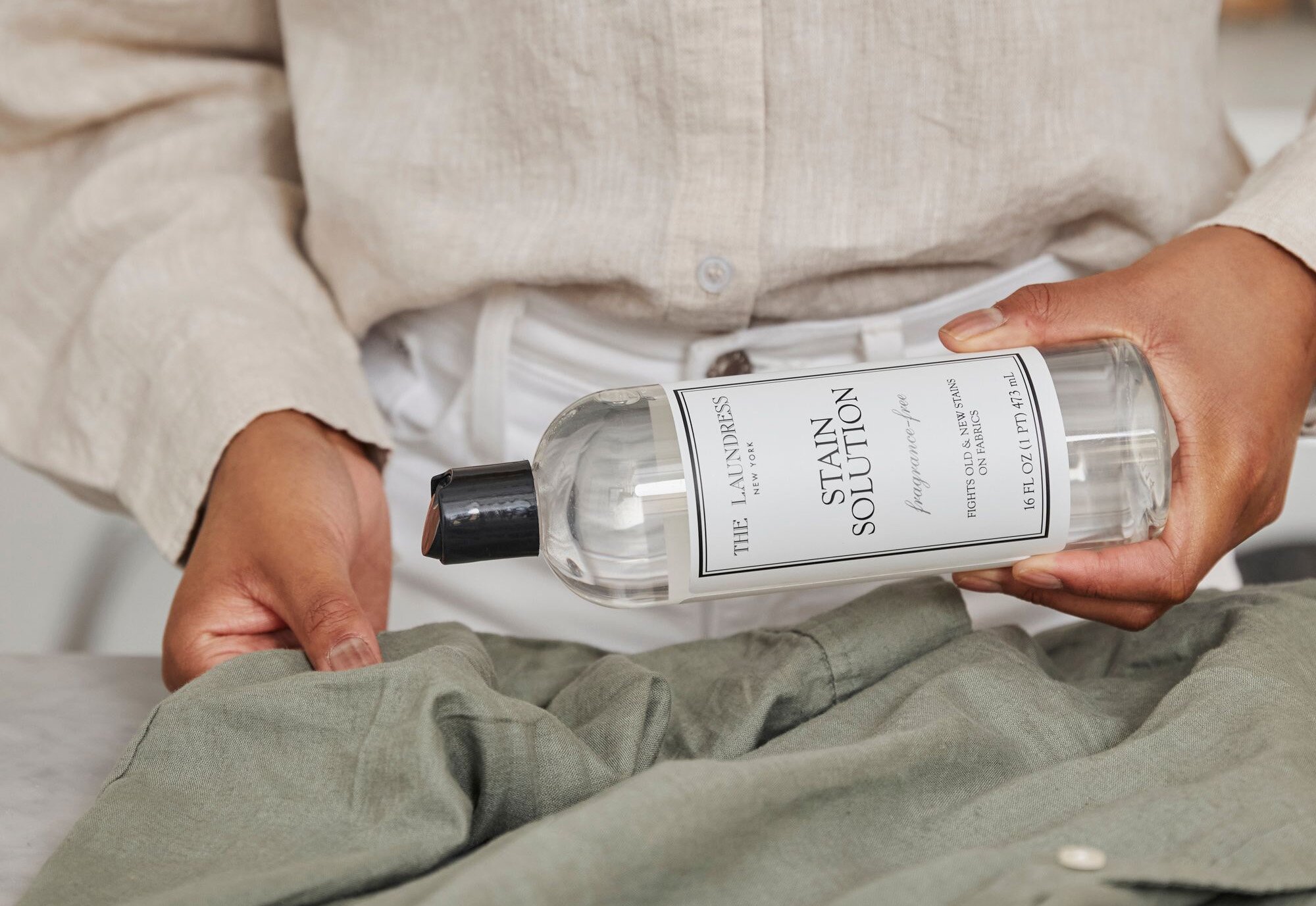 A person wearing white and beige clothing holds a bottle of The Laundress Stain Solution above a green fabric.