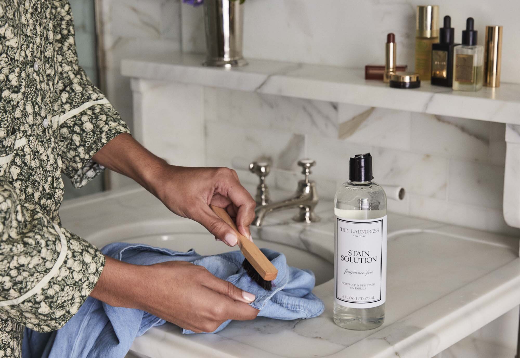 The Laundress Stain Solution sitting on a bathroom countertop beside a lipstick-stained towel and a closed lipstick tube, with a person washing hands under a faucet in a marble sink.