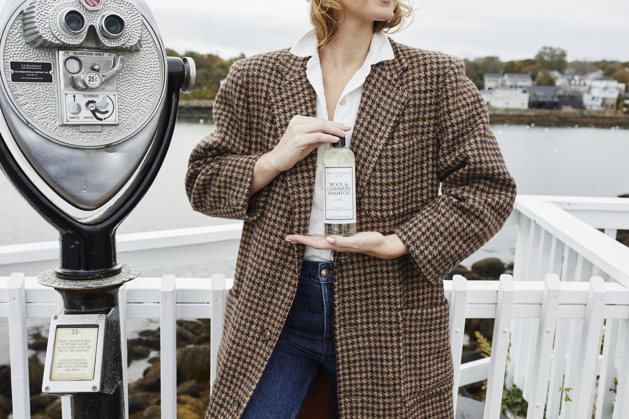 A person in a brown plaid coat holds a bottle of The Laundress Wool & Cashmere Shampoo beside a coin-operated viewer on a waterfront, with houses visible across the water.