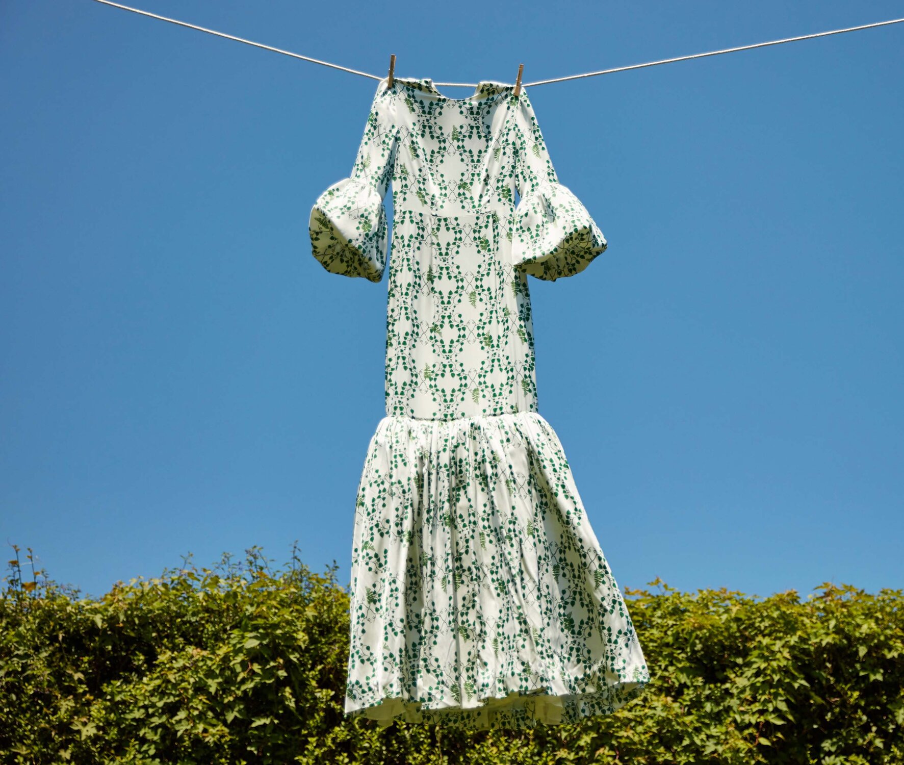 A white dress with green floral patterns hangs on a clothesline, drying under a clear blue sky, after being washed with The Laundress premium fabric care products.