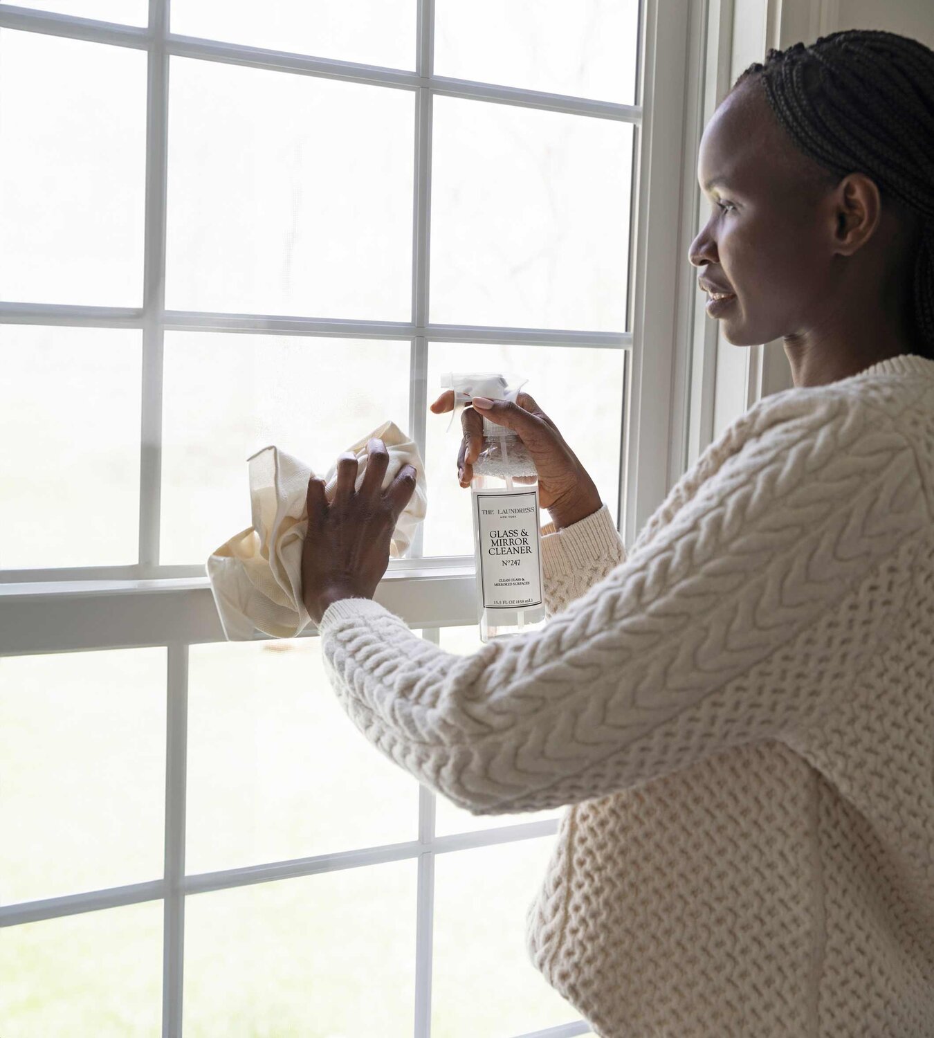 A person in a white sweater cleans a window with a cloth and The Laundress Glass & Mirror Cleaner. The window has multiple panes with a bright, outdoor background.