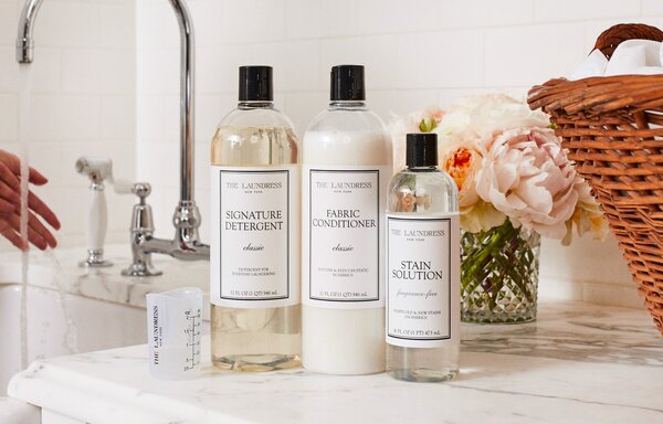 A person washes their hands at a sink, next to which is the laundress signature detergent, fabric conditioner, stain solution, and measuring cup next to a beautiful vase of pale pink flowers