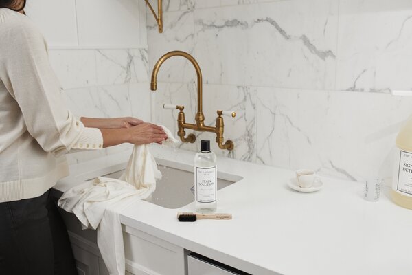 person rinsing coffee stain from white shirt over sink with the laundress stain solution and brush nearby