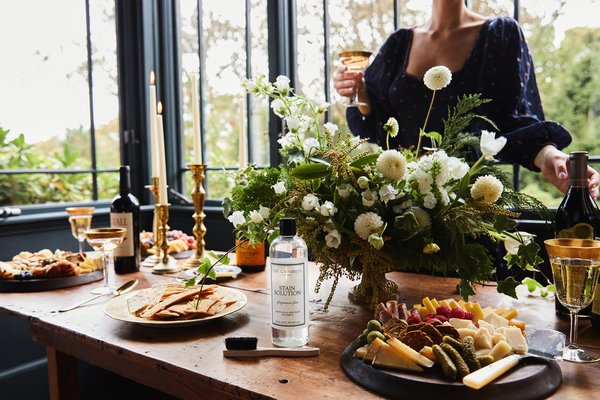 A wooden table holds The Laundress Stain Solution, a floral arrangement, plates of food, and golden candleholders, while a person toasts with a glass of wine in a bright room.