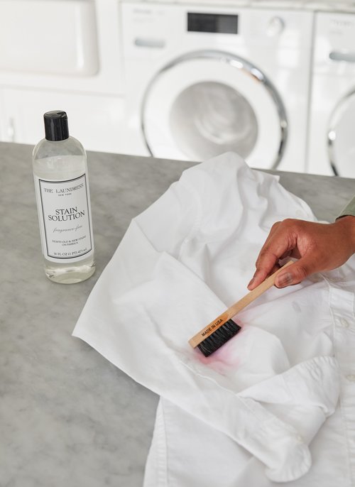A person uses The Laundress fragrance-free Stain Solution and The Laundress Stain Brush to remove a stain from a white garment.