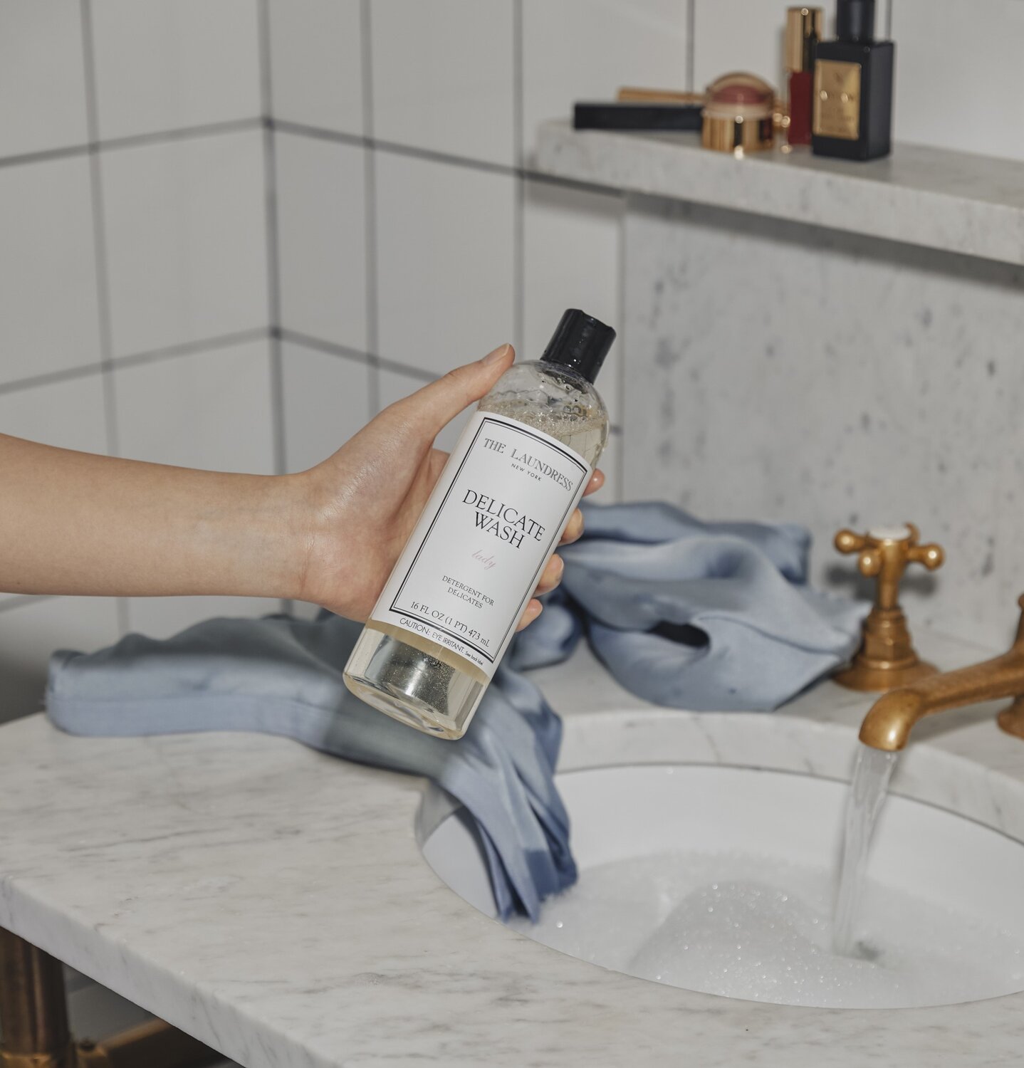 The Laundress Delicate Wash being held over a bathroom sink with running water, with blue garments and toiletries placed on the marble countertop.