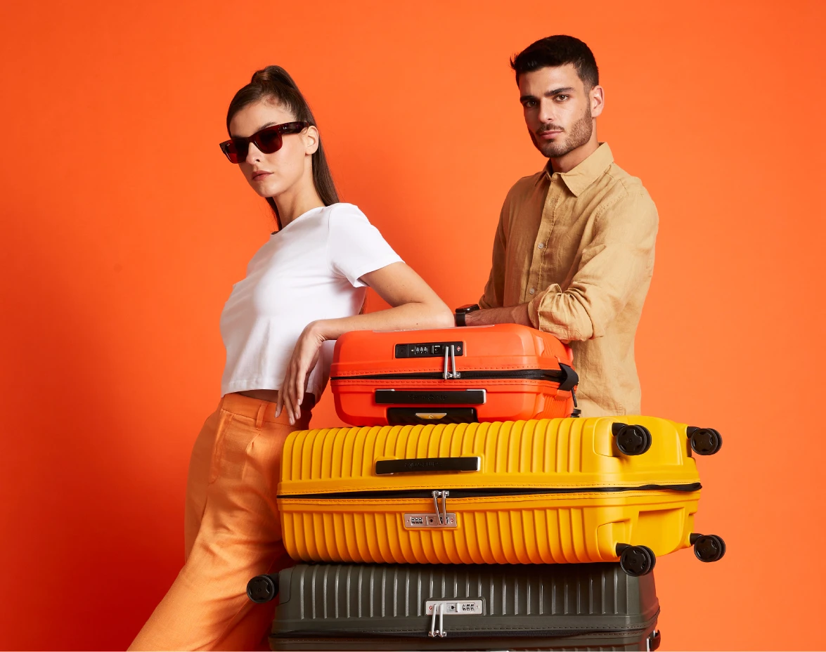 pareja con torre de maletas samsonite de colores vibrantes naranja amarillo y negro fondo naranja