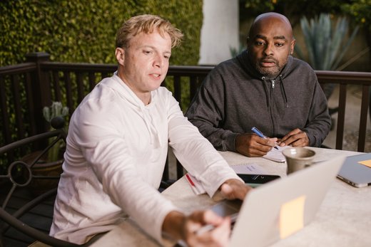 two men outdoors, one in a white jacket and the other in a gray jacket, both deeply engrossed in the laptop screens working together 