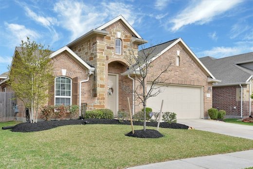 brick wall home with a large closed garage door. In front, there's a lush green front yard, basking in the warmth of a sunny day