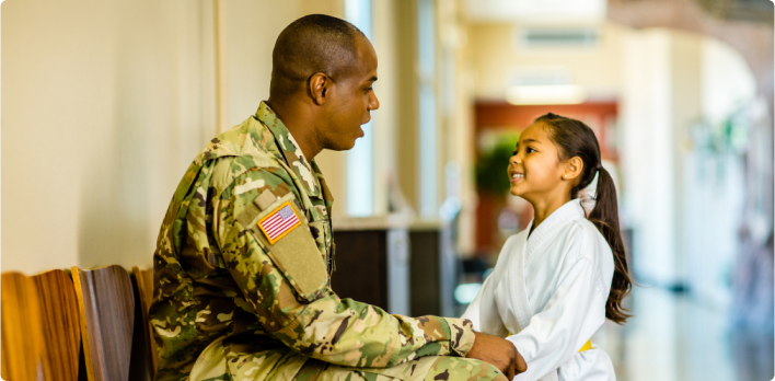 Seated man in US military uniform speaking and holding hands with a smiling young girl in a martial arts outfit. 