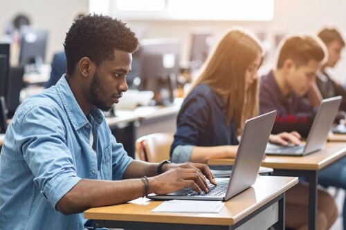 A young man in a light blue denim shirt typing on a laptop at a desk in a classroom or computer lab, with other students working on laptops in the background.