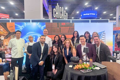 TAO Group Hospitality's backlit display behind a group of smiling professionals. They are wearing business attire and a Chicago sign is seen in the background. There is a table of food in front of the group of people.