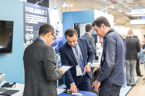 Two men pointing at something on a trade show table in business attire. There is a display behind them and a TV monitor seen to the left.