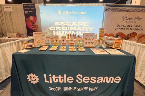 Little Sesame hummus brand booth with teal table cover displaying product samples on plates, branded backdrop reading 'Escape Ordinary Hummus' on sky-blue background, flanked by neighboring vendor displays
