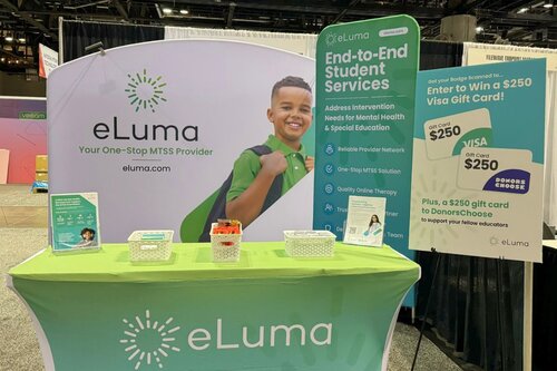 eLuma education services booth with bright lime green branding, curved tension fabric backdrop showing student portrait and 'End-to-End Student Services' messaging, matching table cover, giveaway signage, and organized table display with branded materials