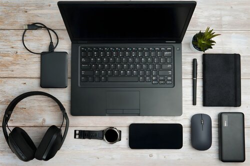 Flat lay of technology desk setup featuring laptop, wireless headphones, external hard drive, mouse, smartphone, smartwatch, notebook and pen on wooden surface
