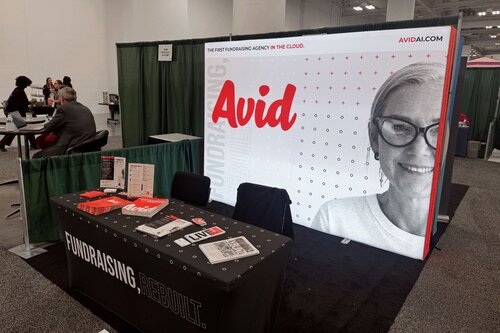 Avid fundraising agency 10x10 trade show booth featuring a large backlit tension fabric display with red branding, black-and-white portrait, and tagline 'The first fundraising agency in the cloud,' alongside a branded table cover reading 'Fundraising Rebuilt' with marketing materials