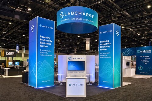 LabCharge trade show booth maximizing vertical space with two tall backlit pillars flanking center display, circular hanging sign reading 'LabCharge: Automate, Integrate, Accelerate,' and branded podium with monitor, all in gradient blue branding