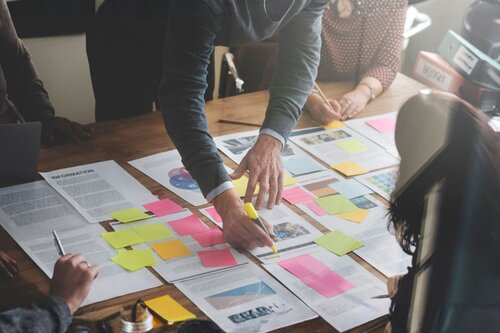 An overhead view of a team planning session with documents, printed reports, and colorful sticky notes spread across a wooden table, with multiple people pointing and writing.