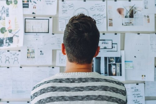Person viewed from behind, studying a wall covered in design sketches, wireframes, and planning documents.