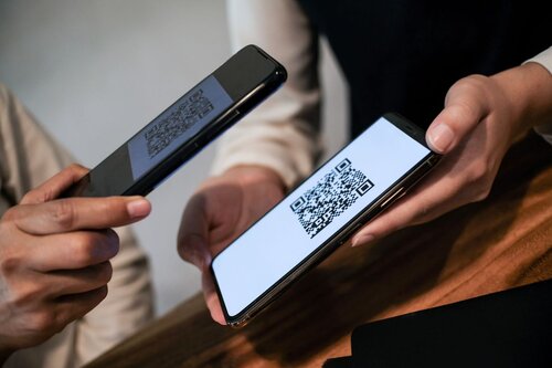 Two hands holding smartphones — one displaying a large QR code on a white screen, the other using its camera to scan it. The exchange takes place over a wooden surface, suggesting a payment or contact-sharing interaction.