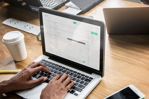 Hands typing on a laptop displaying an email inbox, with a coffee cup and phone on the desk beside it.