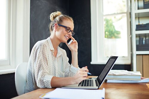 A smiling woman with glasses and a blond bun sits at a wooden desk in a bright home office, holding a smartphone to her ear while typing on a laptop and holding a pen, with stacked papers and documents nearby.