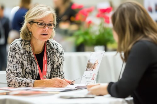 Two people sitting across from each other at a trade show having a conversation at a table.