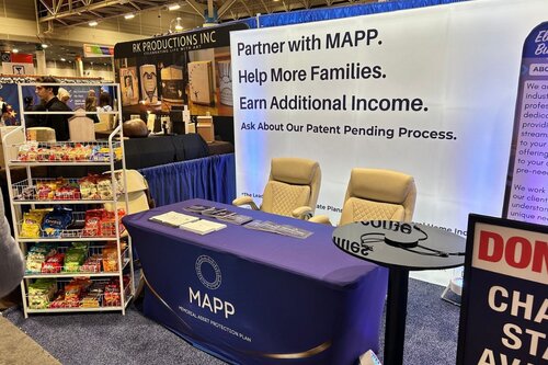 MAPP's trade show booth with a custom display behind two tan chairs positioned at a table. The table has the MAPP logo on it and is dark blue with a circular black podium next to it. There is a snack stand on the far side of the blue table with a variety of snacks.