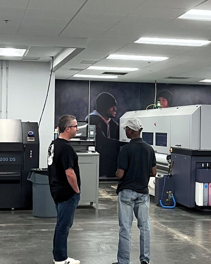 Two be talking to each other facing away from the camera in black shirts. Scott Andrews, the founder of Ace Displays, is on the left and an Ace Connect attendee is on the right. Printing machines are seen in the background.