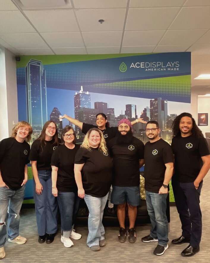 The Ace Displays team posing and smiling for a photo in branded black shirts. A graphic wall with a cityscape is seen behind the group of people.