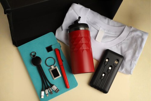 Collection of promotional swag items including teal notebook with keychains, red insulated tumbler, white t-shirt, red pen, and multi-port charging cable