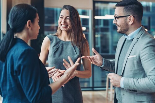 Two women and a man in business attire stand in a bright, glass-walled office, actively conversing with open hand gestures. The woman in the center smiles broadly while listening, conveying engagement and positive energy.