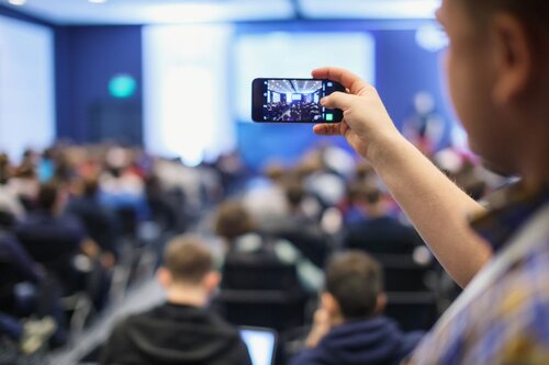 Attendee recording conference presentation on smartphone with large seated audience and illuminated stage in background