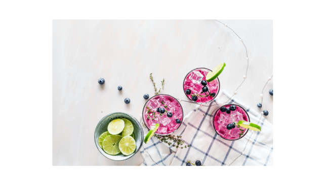 Tres bebidas rosas con hielo, arándanos y rodajas de lima, decoradas con hierbas, sobre una mesa clara.
